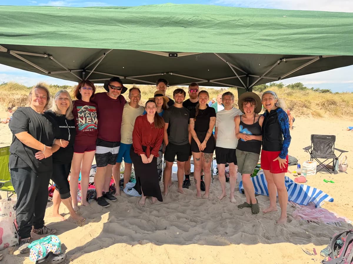 Fifteen people standing together under a gazebo on a sandy beach on a sunny day