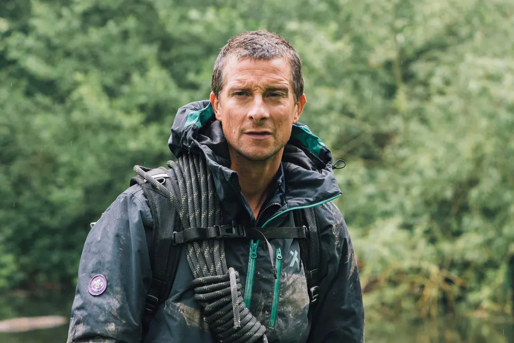 Bear Grylls in a muddy waterproof jacket carrying a coil of rope, standing by a river